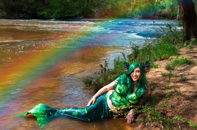 Mermaid in a green tail resting on a riverbank under a rainbow